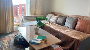 A living room with a sofa, a coffee table with books and a plant, and a view of buildings through a window. - #164 Coral retreat in Birmingham CC -Sleeps 6 (West Midlands)