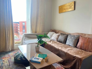 A living room with a sofa, a coffee table with books and a plant, and a view of buildings through a window. - #164 Coral retreat in Birmingham CC -Sleeps 6 (West Midlands)