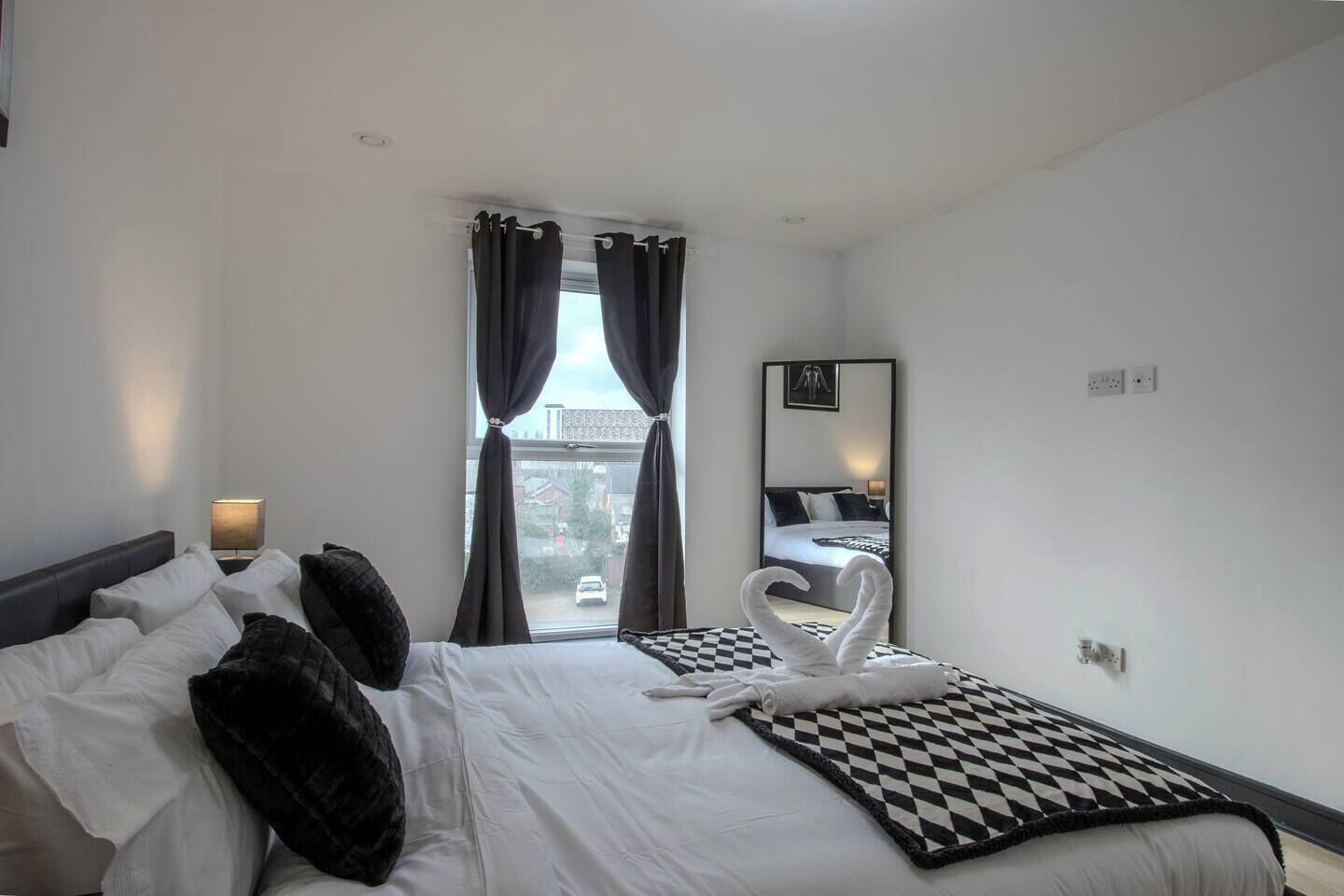 Habitació d'hotel amb un llit, una manta de quadres, una tovallola amb forma de cigne, una làmpada i una finestra amb cortines.