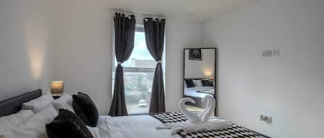 Habitación de hotel con cama, una manta a cuadros, una toalla con forma de cisne, una lámpara y un ventanal con cortinas.