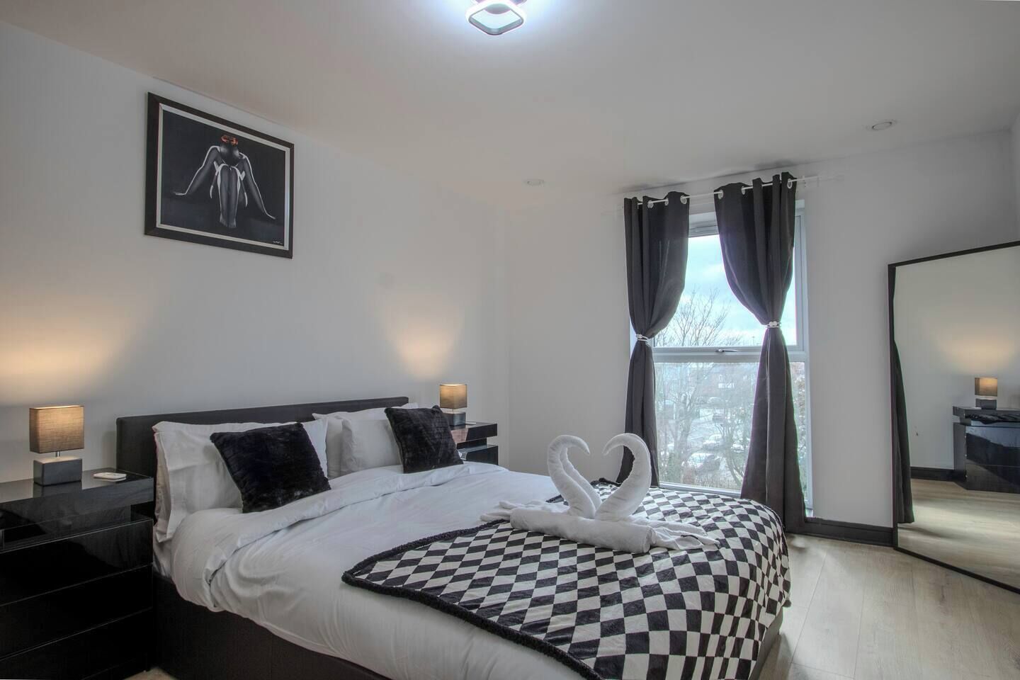 A modern bedroom with a bed, two bedside lamps, a checkered blanket, a heart-shaped towel arrangement, a framed artwork, and a window with curtains.