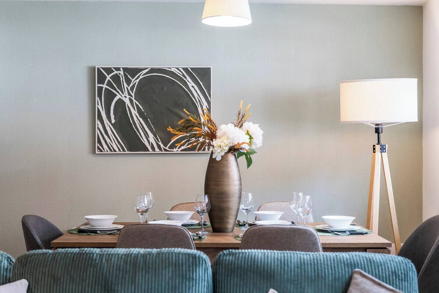 A dining area with a table set for four, a vase with flowers, a floor lamp, and a piece of abstract art on the wall.