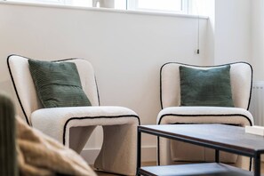 Two chairs with a white and black frame, a green pillow, and a small table in a room with a white wall and a window.