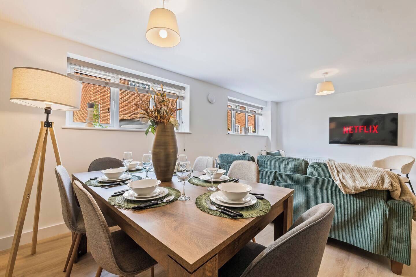 A dining area with a wooden table set for four, a vase with dried plants, and a television displaying 'NETFLIX' in the background.