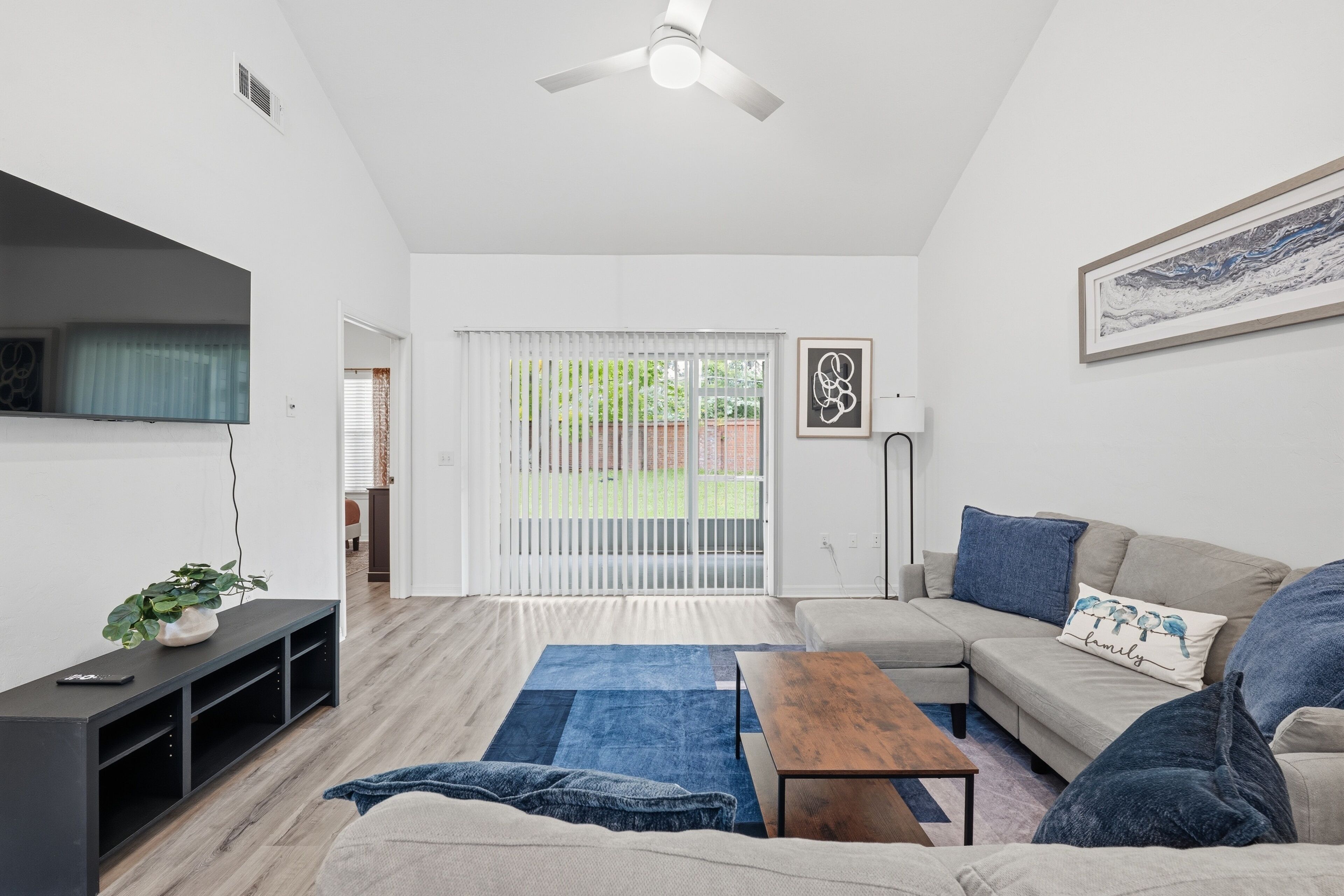 A modern living room with a sofa, coffee table, and a TV on a stand. There is a sliding glass door leading to an outdoor area.