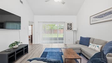 A modern living room with a sofa, coffee table, and a TV on a stand. There is a sliding glass door leading to an outdoor area.