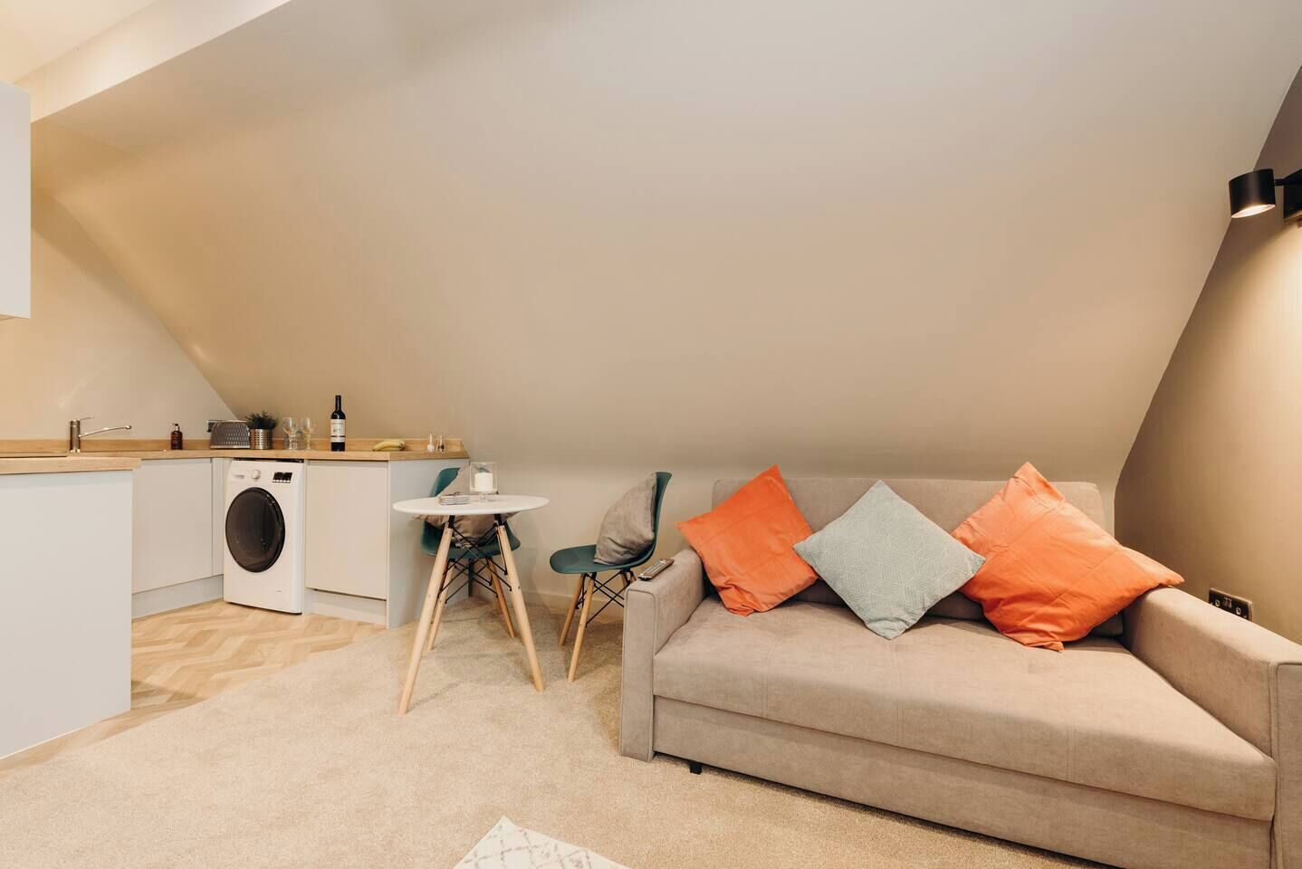 Un espacio habitable compacto con kitchenette, un sofá con cojines, una pequeña mesa y una lavadora.