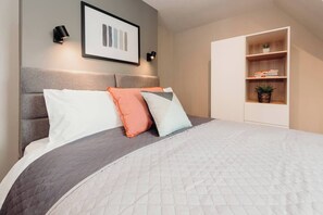 Een netjes opgemaakt bed met een grijs hoofdeinde, witte en oranje kussens en een gewatteerde witte dekbedsprei.