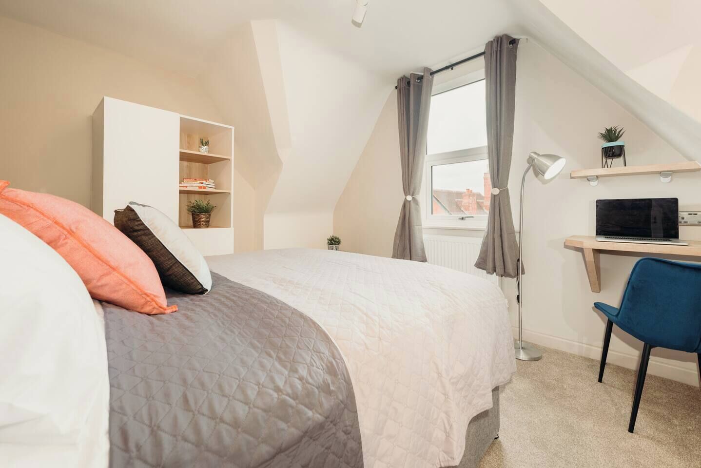 Ein Schlafzimmer mit einem Bett, einem Schreibtisch, einem Stuhl, einem Regal mit Büchern und Pflanzen sowie einem Fenster mit Vorhängen.