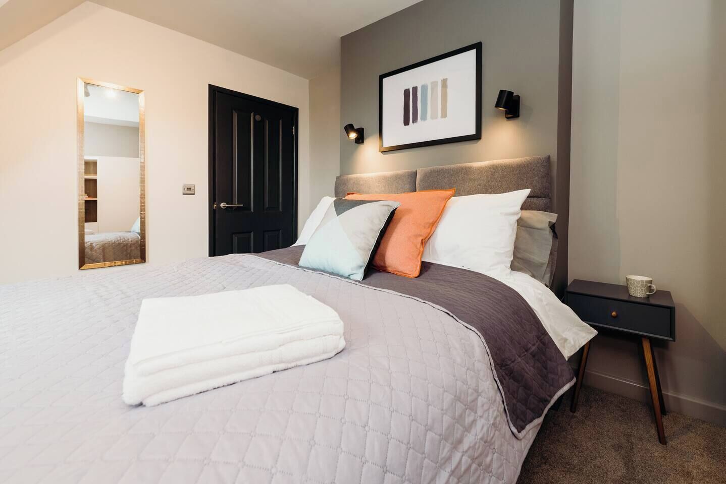 A neatly made bed with a white blanket, a headboard with multiple pillows, a framed picture on the wall, and a bedside table with a cup.