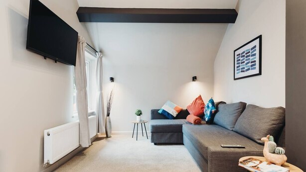 A modern living room with a grey sofa, a flat-screen TV mounted on the wall, a small side table with a book, and a radiator on the floor.