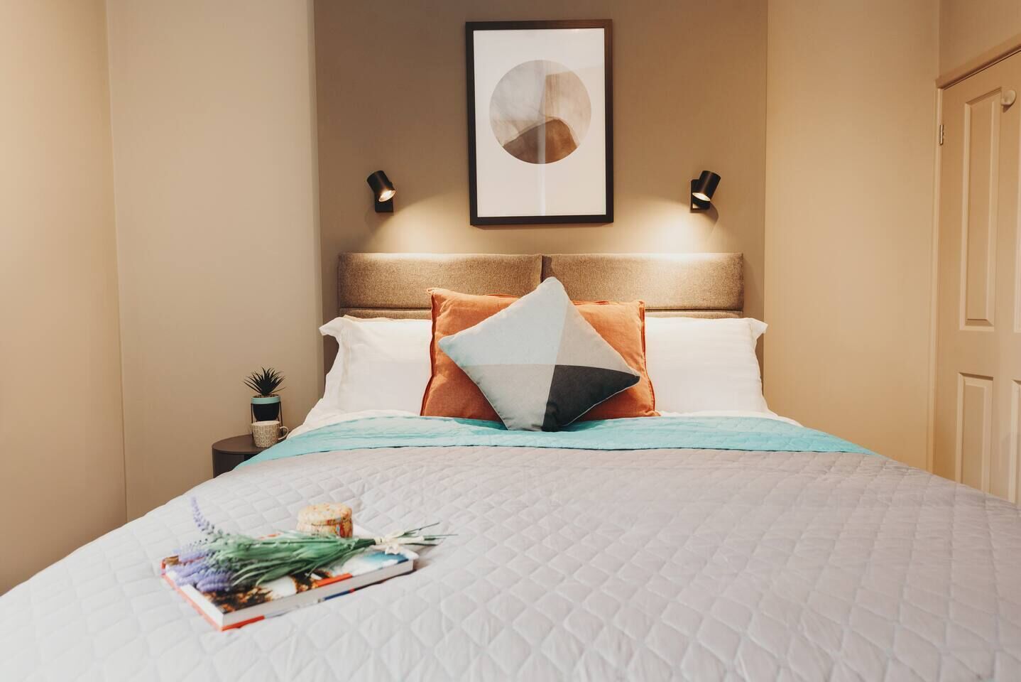Un lit bien fait, recouvert d’une courtepointe matelassée, avec un tableau encadré au-dessus et deux lampes fixées au mur.