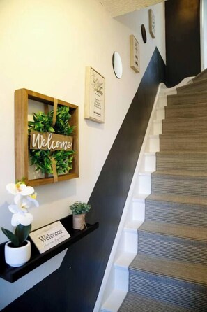 A staircase with a dark handrail and carpeted steps, a welcome sign, and a shelf with a small plant and a welcome card. - Coventry Home: Playroom & CBS Arena (West Midlands)