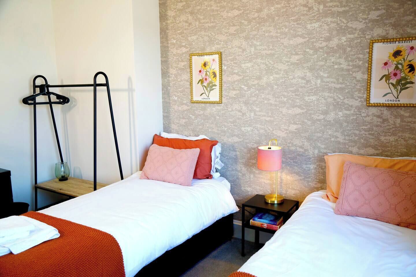 A hotel room with two beds, a bedside table with a lamp, a wall-mounted shelf, and a framed picture of flowers.
