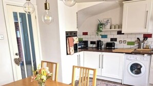 Ein Essbereich mit Holztisch und Stühlen, einer Vase mit Blumen sowie eine Küche im Hintergrund mit weißen Schränken und Geräten.
