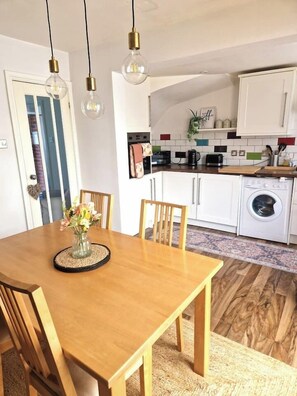 A dining area with a wooden table and chairs, a vase with flowers, and a kitchen in the background with white cabinets and appliances. - Coventry Home: Playroom & CBS Arena (West Midlands)