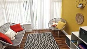 Ein Zimmer mit einem Korbssessel, einem Sessel, einem gemusterten Teppich und einem Bücherregal.