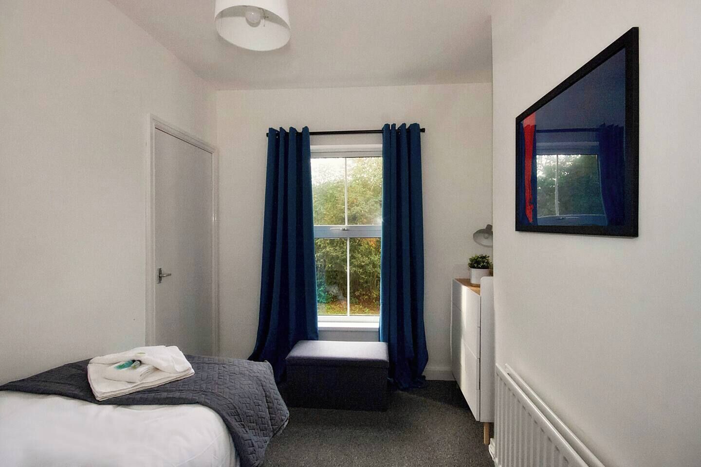 Ein Schlafzimmer mit einem Bett, einem Fenster mit Vorhängen, einem Heizkörper und einem gerahmten Bild an der Wand.