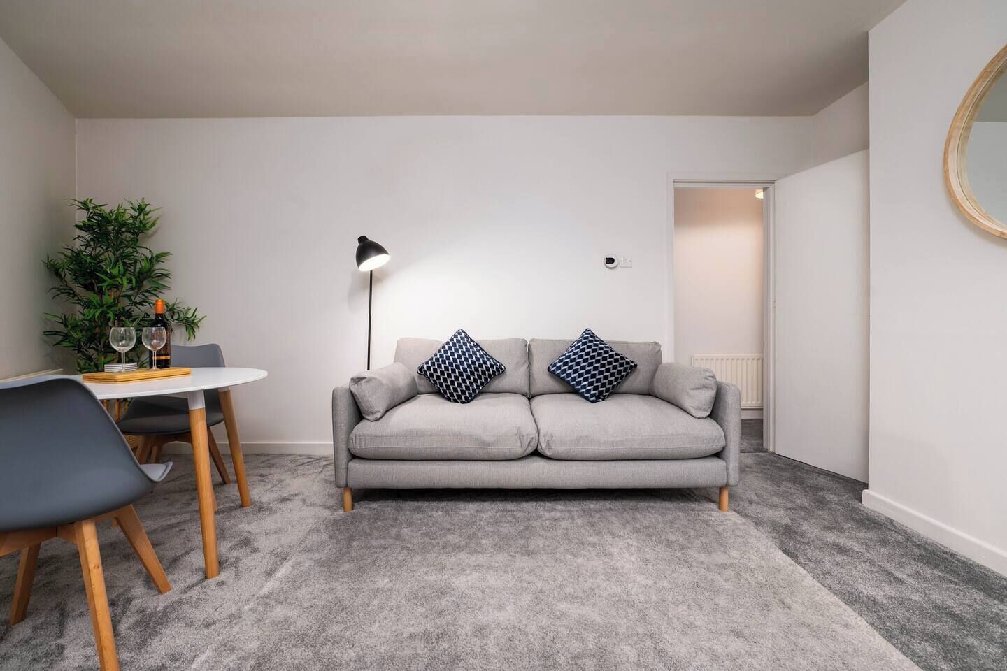 Una sala de estar moderna con un sofá gris, dos cojines estampados, una pequeña mesa redonda con una botella de vino y vasos, una lámpara de pie y una planta en maceta.