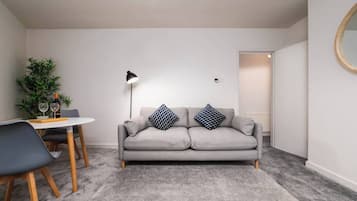 Una sala de estar moderna con un sofá gris, dos cojines estampados, una pequeña mesa redonda con una botella de vino y vasos, una lámpara de pie y una planta en maceta.
