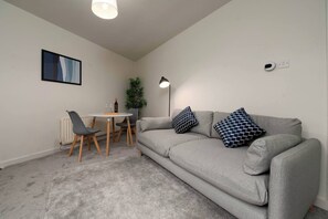 A living room with a grey sofa, two patterned cushions, a small round table with chairs, a floor lamp, a radiator, and a wall-mounted light fixture.