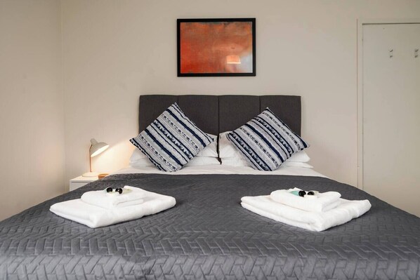 A neatly made bed with a grey quilt, two pillows with a striped pattern, and a bedside lamp.