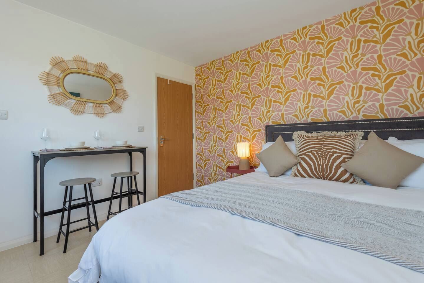A bedroom with a bed, bedside table, lamp, and a desk with two stools.