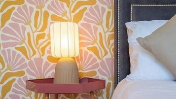 A bedroom with a patterned wallpaper, a bedside lamp, a bed with pillows, and a wooden floor.