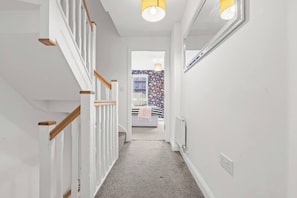 Un pasillo con una escalera, un radiador y una puerta que da a una habitación con cama y ventana.