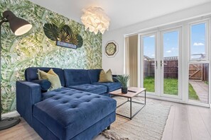 Una sala de estar con un sofá seccional azul, una mesita, una lámpara de pared y un ventanal con vistas a un jardín.