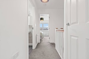 Un pasillo con piso alfombrado, paredes blancas y una puerta que da a un dormitorio con ventana.