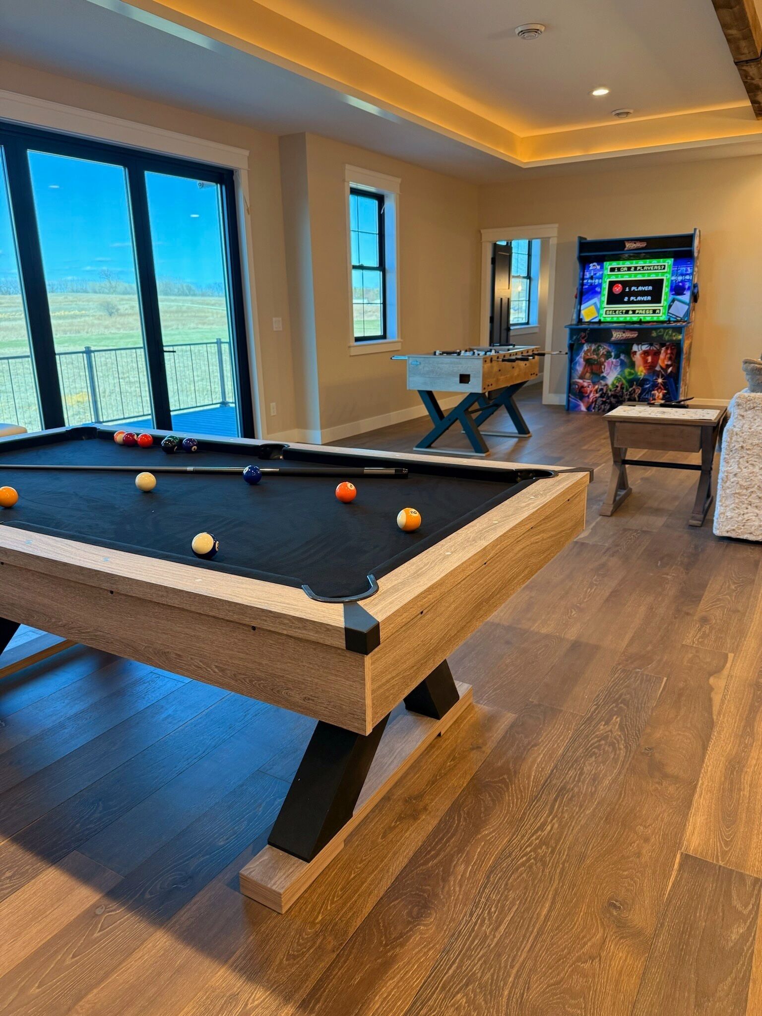 Ein Billardtisch mit Kugeln, ein Kicker und eine Spielkonsole in einem Raum mit großen Fenstern und Blick ins Freie.