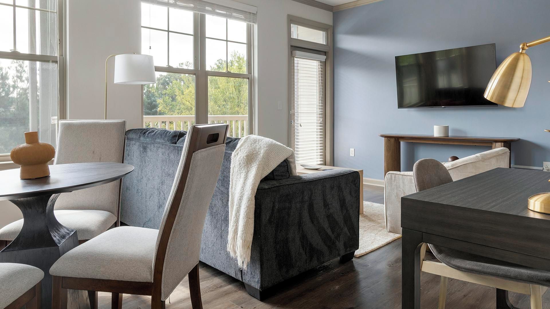 Una sala moderna con una mesa de comedor de madera oscura, sillas de colores claros, un televisor plano grande y un ventanal con persianas.
