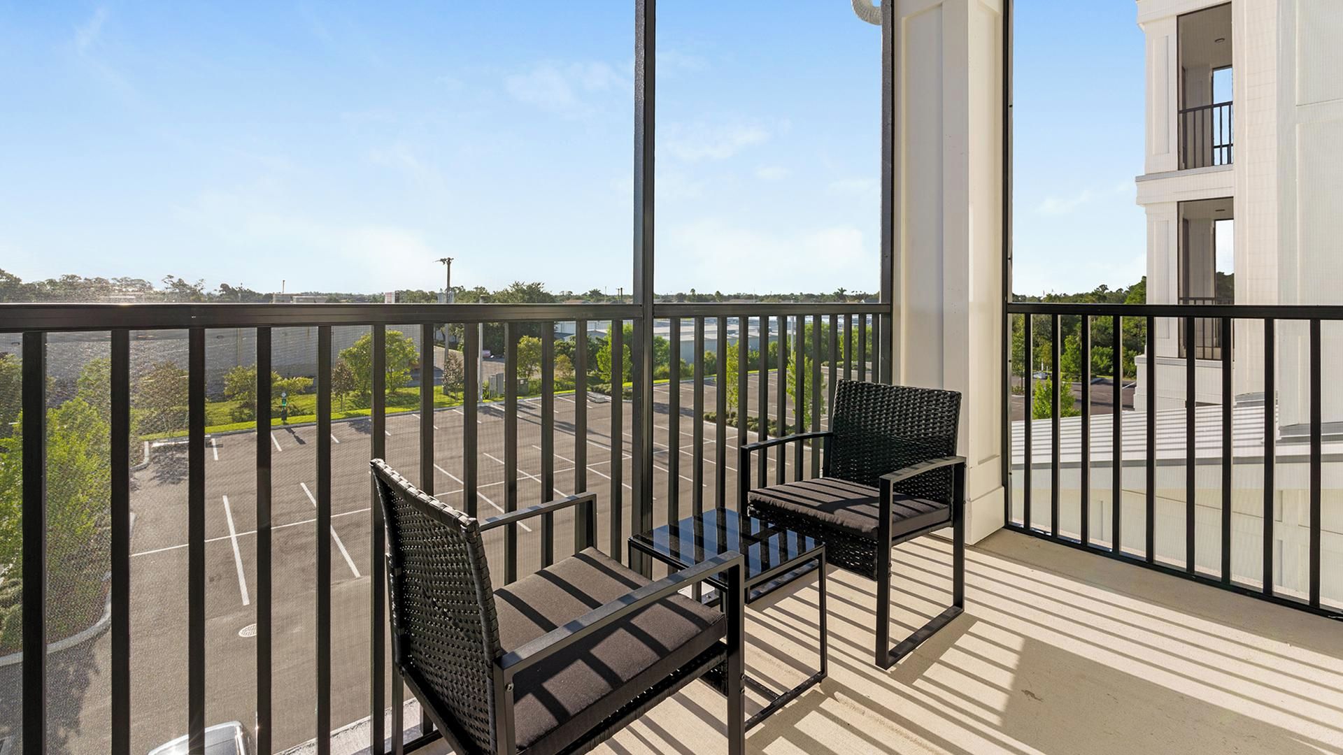 A balcony with two chairs and a table, overlooking a parking lot and greenery.