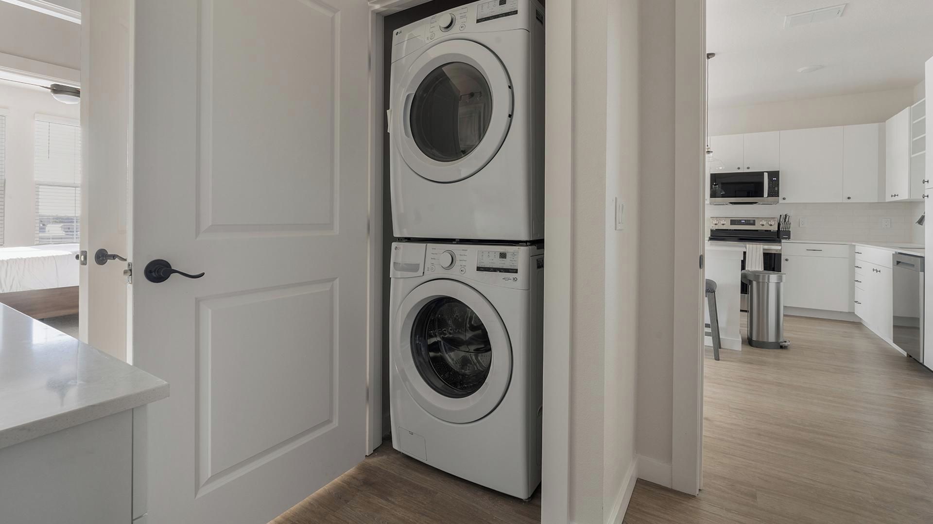 A laundry room with two stacked washing machines, a kitchen area with modern appliances, and a view of the outside through an open door.