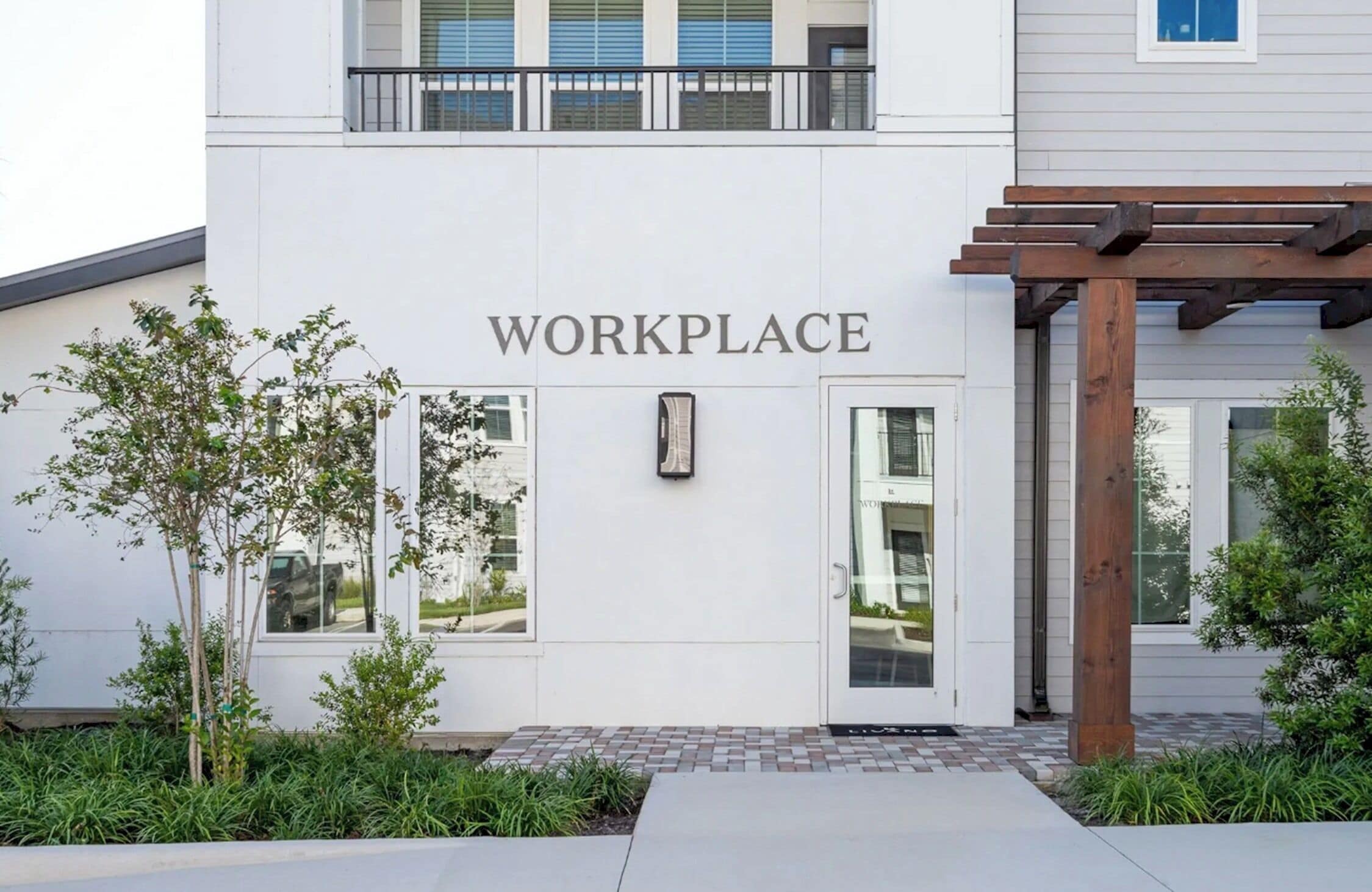 A building with a sign reading 'WORKPLACE' above the entrance, a wooden pergola, and a small tree in the foreground.