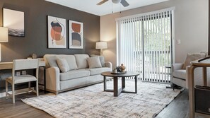 Ein Wohnzimmer mit einer Couch, einem Couchtisch, einem Schreibtisch und einem Stuhl. An der Wand befinden sich zwei abstrakte Gemälde, ein Deckenventilator und ein Fenster mit Jalousien.