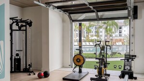 Ett gym med träningsutrustning, en fläkt och utsikt över en swimmingpool och byggnader.