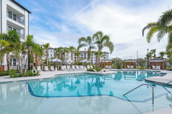 A swimming pool with lounge chairs and umbrellas, surrounded by palm trees and buildings. - Landing Beautiful 1bd, Gym, Clubhouse (Port Charlotte)