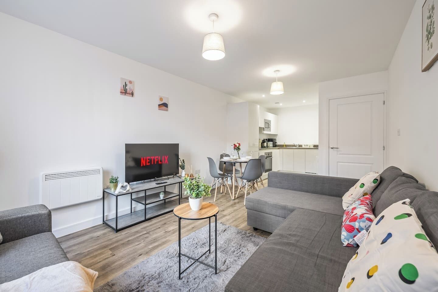 A modern living room with a flat-screen TV, grey sofas, a dining area with chairs, a small table with a plant, and a kitchen in the background.