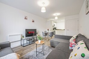 Ein modernes Wohnzimmer mit Flachbildfernseher, grauen Sofas, einer Essecke mit Stühlen, einem kleinen Tisch mit einer Pflanze und einer Küche im Hintergrund.