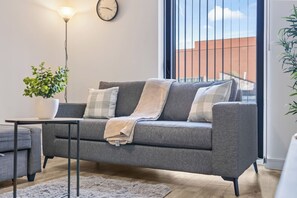 Ett modernt vardagsrum med en grå soffa, ett litet bord, en krukväxt och ett stort fönster med utsikt över byggnader.