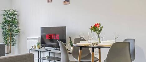A modern living room with a dining area, a flat-screen TV displaying 'NETFLIX,' and a potted plant.