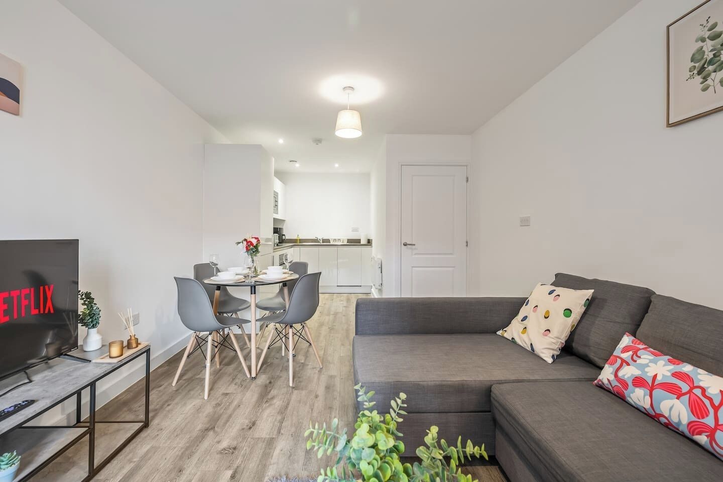 Un salon moderne avec un canapé gris, une table à manger avec des chaises, un téléviseur et une cuisine en arrière-plan.