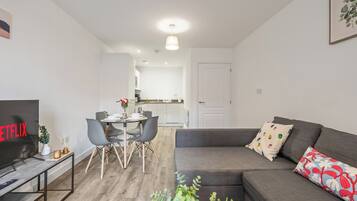 A modern living room with a grey sofa, a dining table with chairs, a TV, and a kitchen in the background.