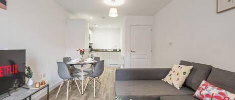 Una sala de estar moderna con un sofá gris, una mesa de comedor con sillas, un televisor y una cocina al fondo.
