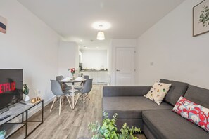 Ein modernes Wohnzimmer mit einem grauen Sofa, einem Esstisch mit Stühlen, einem Fernseher und einer Küche im Hintergrund.