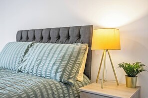 Ein Bett mit einem geknöpften Kopfteil, eine Nachttischlampe mit goldfarbener Basis und ein Blumentopf auf dem Nachttisch.
