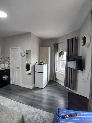 Un espacio habitable compacto con kitchenette, lavadora, refrigerador pequeño, televisor de pantalla plana montado en la pared y una ventana con cortinas.