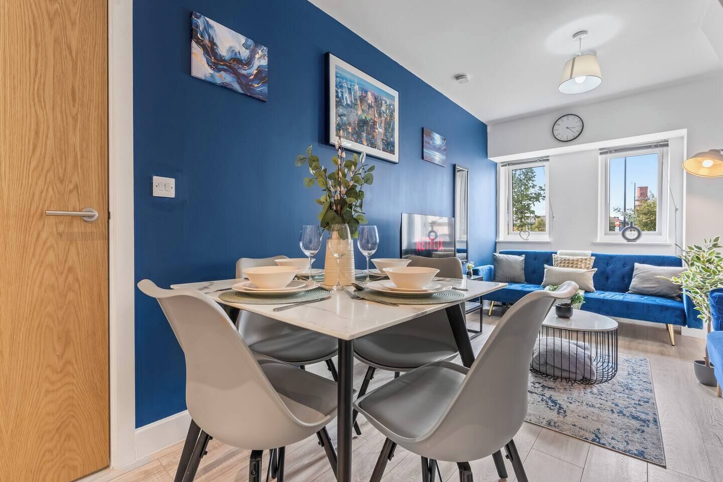 A modern living room with a blue accent wall, a dining area with a table set for two, and a sofa with a view of the outdoors.